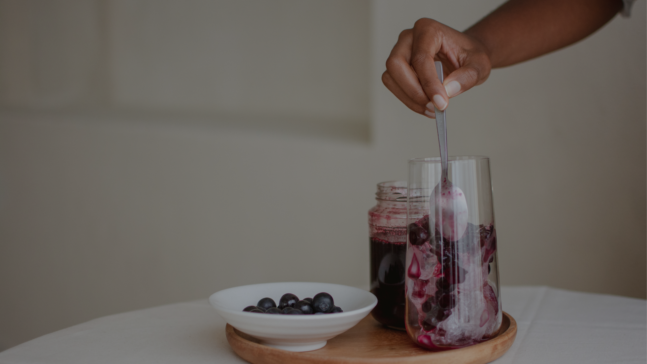 a woman's hand stirring a glass with a purple drink on a table that also has a small bowl of blueberries on it.